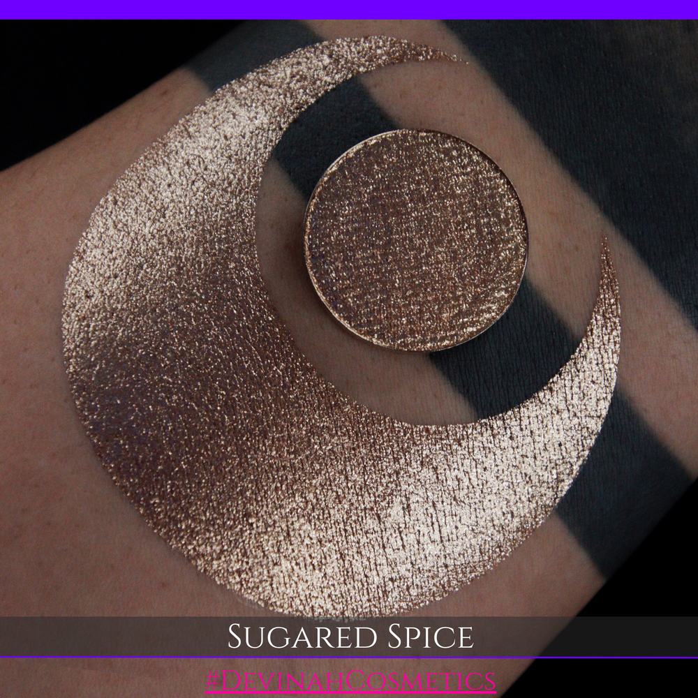 A round eyeshadow pan labeled 'SUGARED SPICE' made by Devinah Cosmetics, placed over a large crescent moon swatch of the same shimmering shifty eyeshadow on skin. The eyeshadow has sparkly colors of beige creme shimmer.