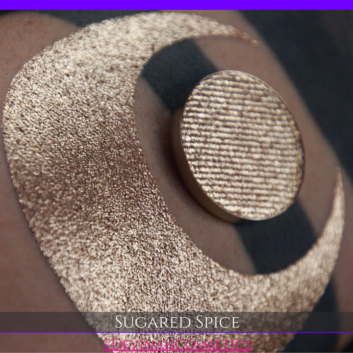 A round eyeshadow pan labeled 'SUGARED SPICE' made by Devinah Cosmetics, placed over a large crescent moon swatch of the same shimmering shifty eyeshadow on skin. The eyeshadow has sparkly colors of beige creme shimmer.