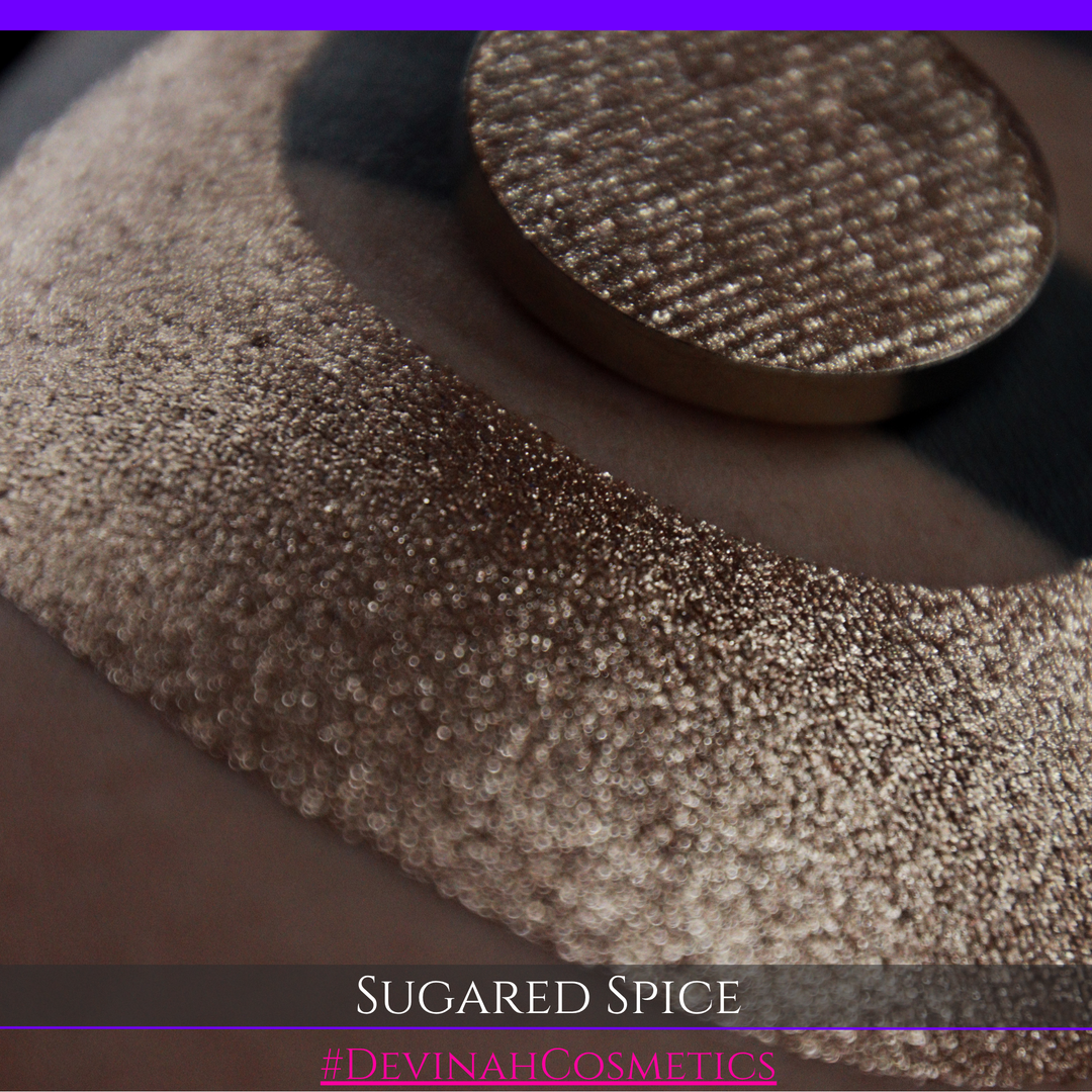 A round eyeshadow pan labeled 'SUGARED SPICE' made by Devinah Cosmetics, placed over a large crescent moon swatch of the same shimmering shifty eyeshadow on skin. The eyeshadow has sparkly colors of beige creme shimmer.