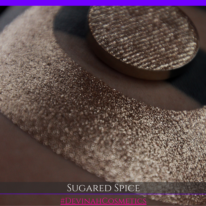 A round eyeshadow pan labeled 'SUGARED SPICE' made by Devinah Cosmetics, placed over a large crescent moon swatch of the same shimmering shifty eyeshadow on skin. The eyeshadow has sparkly colors of beige creme shimmer.