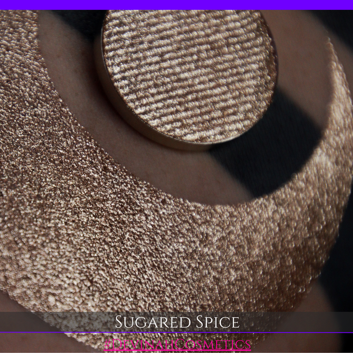 A round eyeshadow pan labeled 'SUGARED SPICE' made by Devinah Cosmetics, placed over a large crescent moon swatch of the same shimmering shifty eyeshadow on skin. The eyeshadow has sparkly colors of beige creme shimmer.