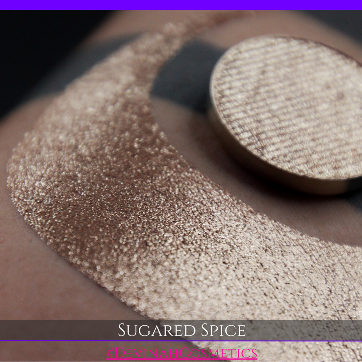 A round eyeshadow pan labeled 'SUGARED SPICE' made by Devinah Cosmetics, placed over a large crescent moon swatch of the same shimmering shifty eyeshadow on skin. The eyeshadow has sparkly colors of beige creme shimmer.