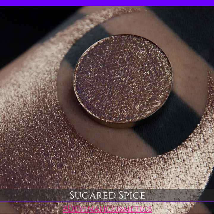 A round eyeshadow pan labeled 'SUGARED SPICE' made by Devinah Cosmetics, placed over a large crescent moon swatch of the same shimmering shifty eyeshadow on skin. The eyeshadow has sparkly colors of beige creme shimmer.