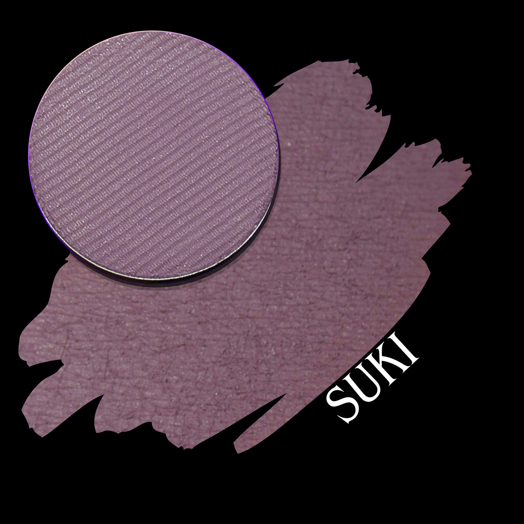 A makeup swatch showing a circular pressed powder eyeshadow pan in a dusty mauve-purple shade on a black background. The word 'SUKI' appears in white near the lower right.