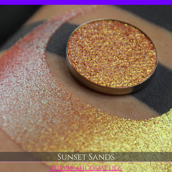 A round eyeshadow pan labeled 'SUNSET SANDS' made by Devinah Cosmetics, placed over a large crescent moon swatch of the same shimmering shifty eyeshadow on skin. The eyeshadow has a duochrome multichrome shift between colors peachy coral pink, orange, yellow, gold.