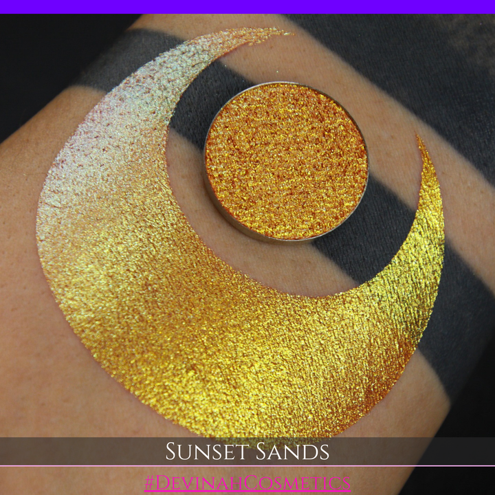 A round eyeshadow pan labeled 'SUNSET SANDS' made by Devinah Cosmetics, placed over a large crescent moon swatch of the same shimmering shifty eyeshadow on skin. The eyeshadow has a duochrome multichrome shift between colors peachy coral pink, orange, yellow, gold.