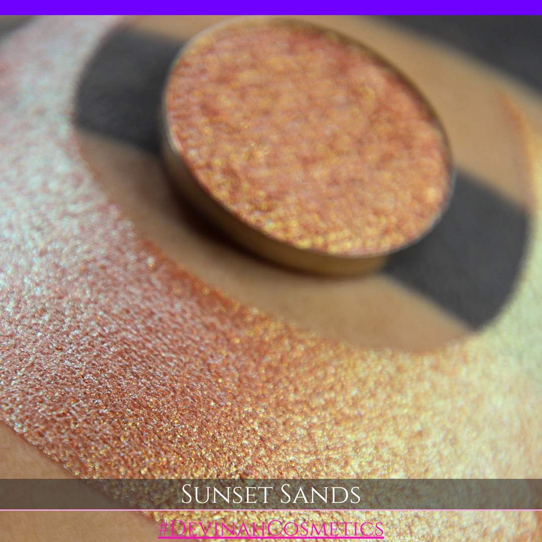A round eyeshadow pan labeled 'SUNSET SANDS' made by Devinah Cosmetics, placed over a large crescent moon swatch of the same shimmering shifty eyeshadow on skin. The eyeshadow has a duochrome multichrome shift between colors peachy coral pink, orange, yellow, gold.