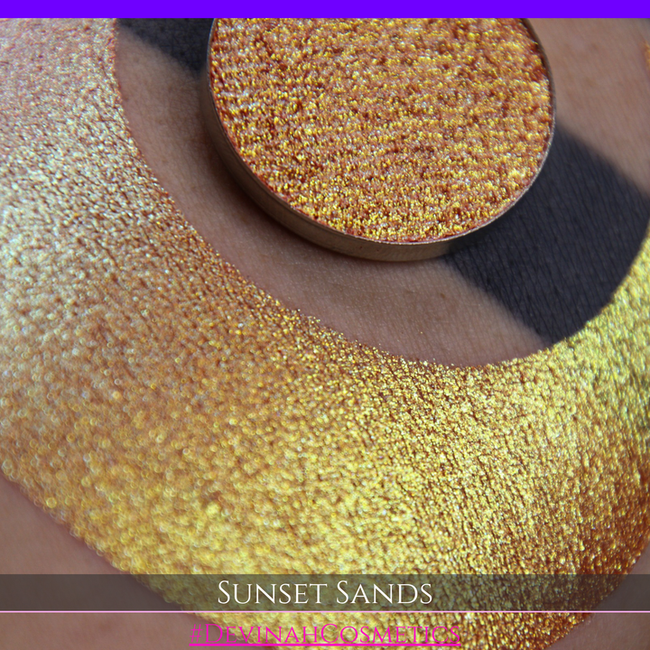 A round eyeshadow pan labeled 'SUNSET SANDS' made by Devinah Cosmetics, placed over a large crescent moon swatch of the same shimmering shifty eyeshadow on skin. The eyeshadow has a duochrome multichrome shift between colors peachy coral pink, orange, yellow, gold.