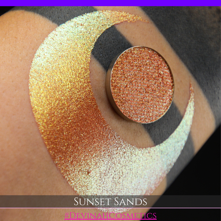 A round eyeshadow pan labeled 'SUNSET SANDS' made by Devinah Cosmetics, placed over a large crescent moon swatch of the same shimmering shifty eyeshadow on skin. The eyeshadow has a duochrome multichrome shift between colors peachy coral pink, orange, yellow, gold.