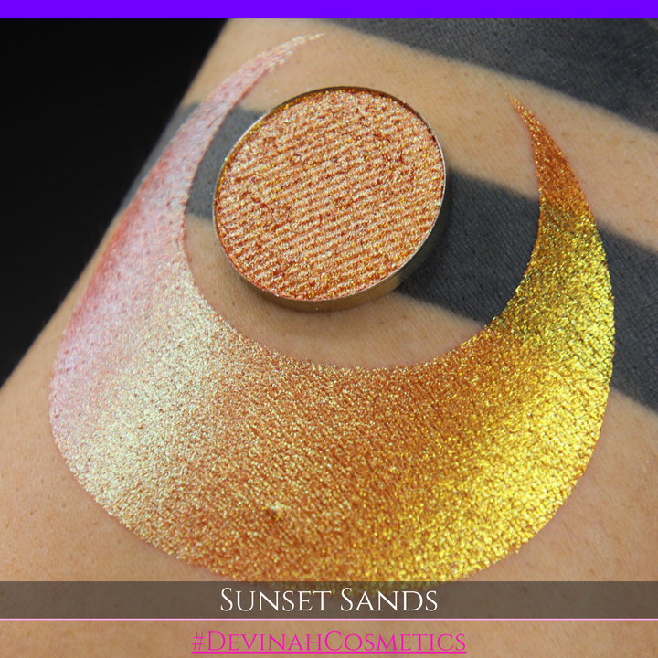 A round eyeshadow pan labeled 'SUNSET SANDS' made by Devinah Cosmetics, placed over a large crescent moon swatch of the same shimmering shifty eyeshadow on skin. The eyeshadow has a duochrome multichrome shift between colors peachy coral pink, orange, yellow, gold.