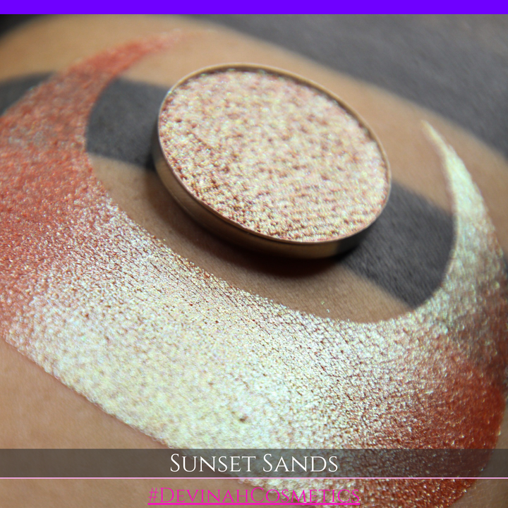 A round eyeshadow pan labeled 'SUNSET SANDS' made by Devinah Cosmetics, placed over a large crescent moon swatch of the same shimmering shifty eyeshadow on skin. The eyeshadow has a duochrome multichrome shift between colors peachy coral pink, orange, yellow, gold.