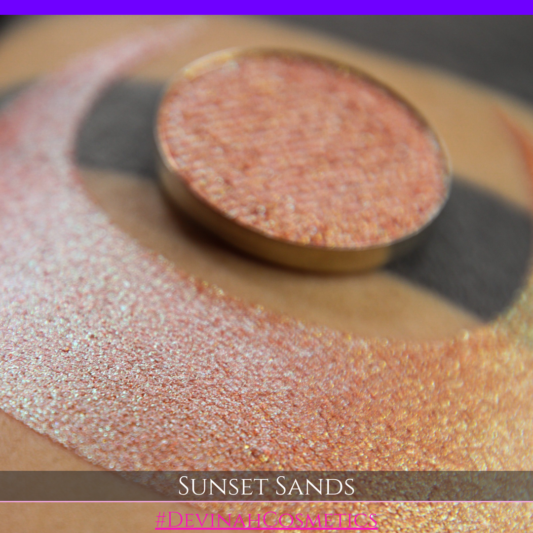 A round eyeshadow pan labeled 'SUNSET SANDS' made by Devinah Cosmetics, placed over a large crescent moon swatch of the same shimmering shifty eyeshadow on skin. The eyeshadow has a duochrome multichrome shift between colors peachy coral pink, orange, yellow, gold.