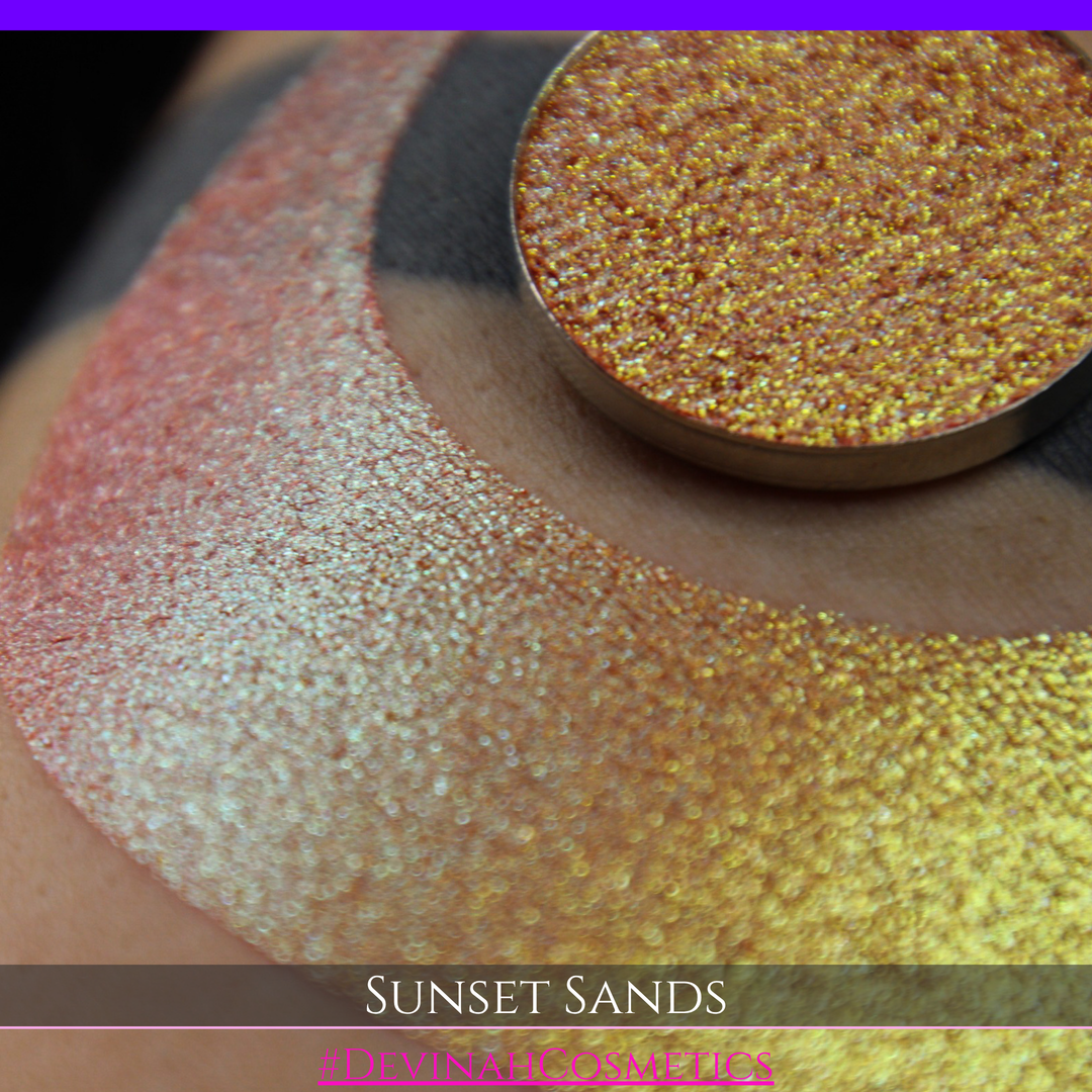 A round eyeshadow pan labeled 'SUNSET SANDS' made by Devinah Cosmetics, placed over a large crescent moon swatch of the same shimmering shifty eyeshadow on skin. The eyeshadow has a duochrome multichrome shift between colors peachy coral pink, orange, yellow, gold.
