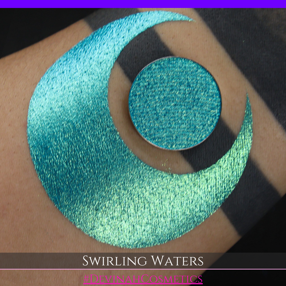 A round eyeshadow pan labeled 'SWIRLING WATERS' made by Devinah Cosmetics, placed over a large crescent moon swatch of the same shimmering shifty eyeshadow on skin. The eyeshadow has a duochrome multichrome shift between colors teal, blue, green.