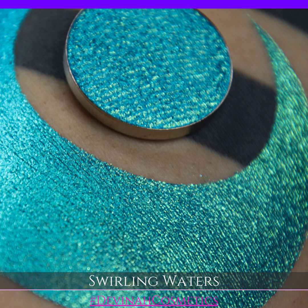A round eyeshadow pan labeled 'SWIRLING WATERS' made by Devinah Cosmetics, placed over a large crescent moon swatch of the same shimmering shifty eyeshadow on skin. The eyeshadow has a duochrome multichrome shift between colors teal, blue, green.