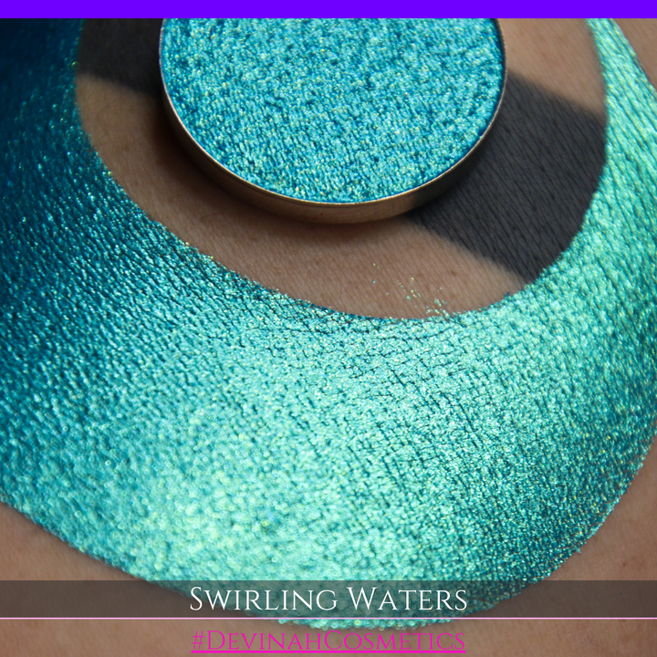 A round eyeshadow pan labeled 'SWIRLING WATERS' made by Devinah Cosmetics, placed over a large crescent moon swatch of the same shimmering shifty eyeshadow on skin. The eyeshadow has a duochrome multichrome shift between colors teal, blue, green.