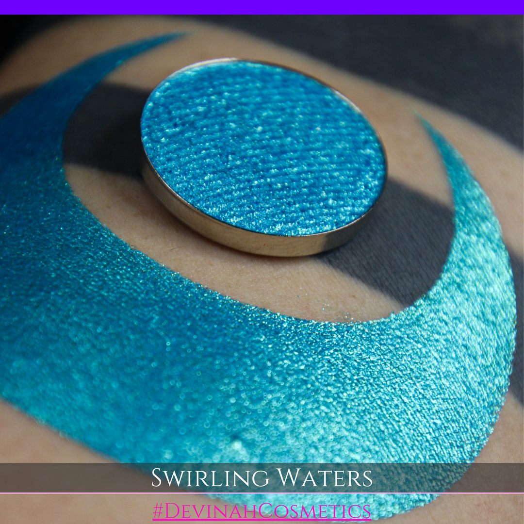 A round eyeshadow pan labeled 'SWIRLING WATERS' made by Devinah Cosmetics, placed over a large crescent moon swatch of the same shimmering shifty eyeshadow on skin. The eyeshadow has a duochrome multichrome shift between colors teal, blue, green.