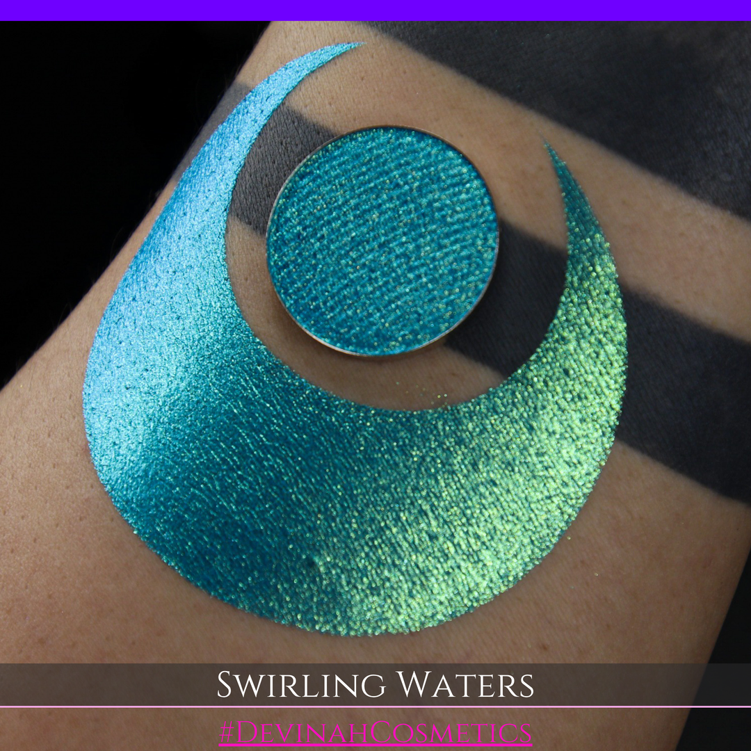 A round eyeshadow pan labeled 'SWIRLING WATERS' made by Devinah Cosmetics, placed over a large crescent moon swatch of the same shimmering shifty eyeshadow on skin. The eyeshadow has a duochrome multichrome shift between colors teal, blue, green.