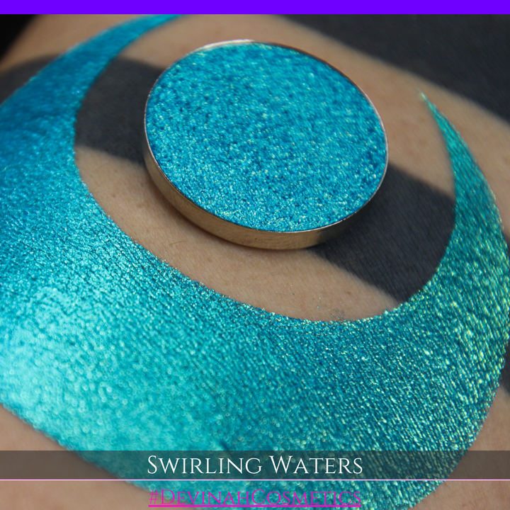 A round eyeshadow pan labeled 'SWIRLING WATERS' made by Devinah Cosmetics, placed over a large crescent moon swatch of the same shimmering shifty eyeshadow on skin. The eyeshadow has a duochrome multichrome shift between colors teal, blue, green.