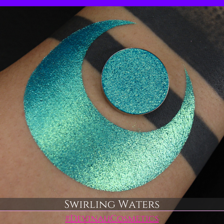 A round eyeshadow pan labeled 'SWIRLING WATERS' made by Devinah Cosmetics, placed over a large crescent moon swatch of the same shimmering shifty eyeshadow on skin. The eyeshadow has a duochrome multichrome shift between colors teal, blue, green.