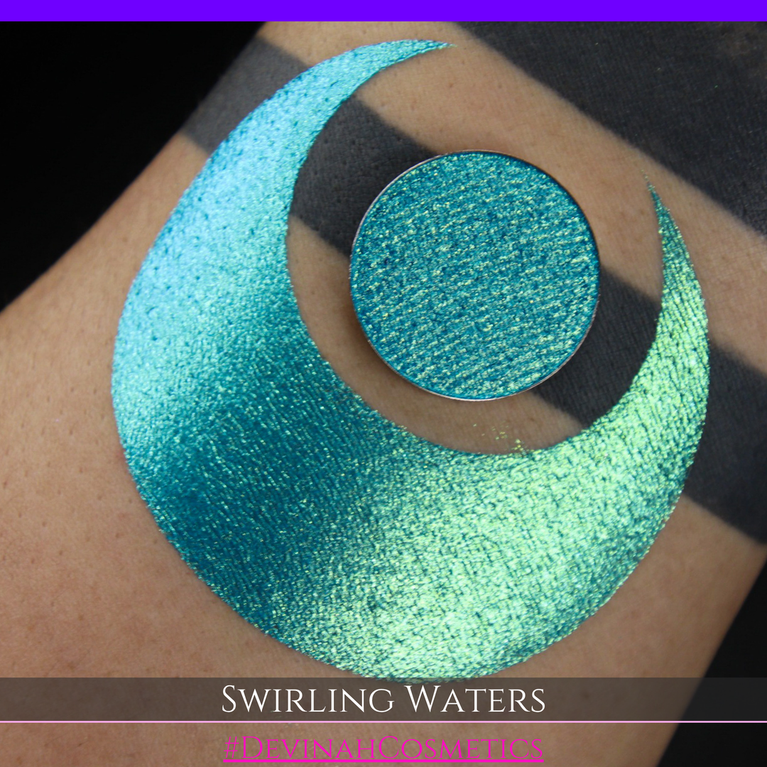 A round eyeshadow pan labeled 'SWIRLING WATERS' made by Devinah Cosmetics, placed over a large crescent moon swatch of the same shimmering shifty eyeshadow on skin. The eyeshadow has a duochrome multichrome shift between colors teal, blue, green.
