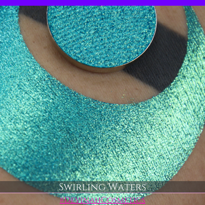 A round eyeshadow pan labeled 'SWIRLING WATERS' made by Devinah Cosmetics, placed over a large crescent moon swatch of the same shimmering shifty eyeshadow on skin. The eyeshadow has a duochrome multichrome shift between colors teal, blue, green.