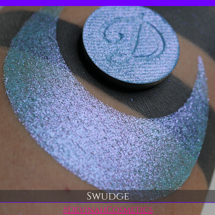 A round eyeshadow pan labeled 'SWUDGE' made by Devinah Cosmetics, placed over a large crescent moon swatch of the same shimmering shifty eyeshadow on skin. The eyeshadow has a duochrome multichrome shift between colors pink, teal, blue.