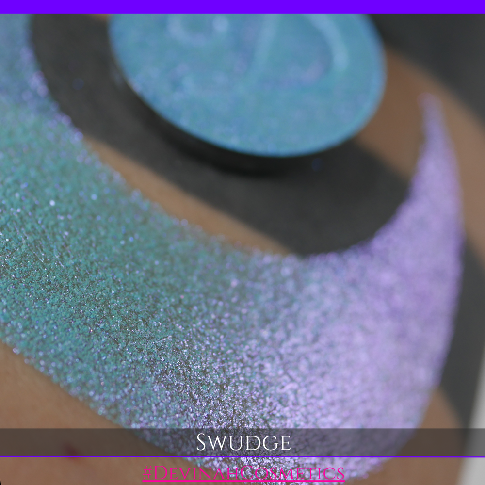 A round eyeshadow pan labeled 'SWUDGE' made by Devinah Cosmetics, placed over a large crescent moon swatch of the same shimmering shifty eyeshadow on skin. The eyeshadow has a duochrome multichrome shift between colors pink, teal, blue.