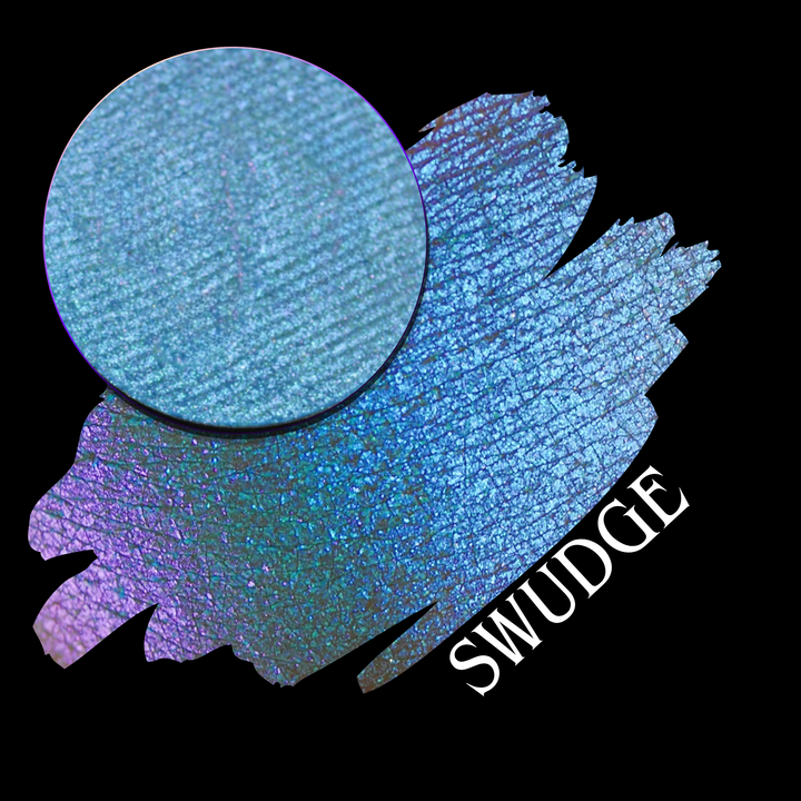 A round eyeshadow pan labeled 'SWUDGE' made by Devinah Cosmetics, placed over a large crescent moon swatch of the same shimmering shifty eyeshadow on skin. The eyeshadow has a duochrome multichrome shift between colors pink, teal, blue.