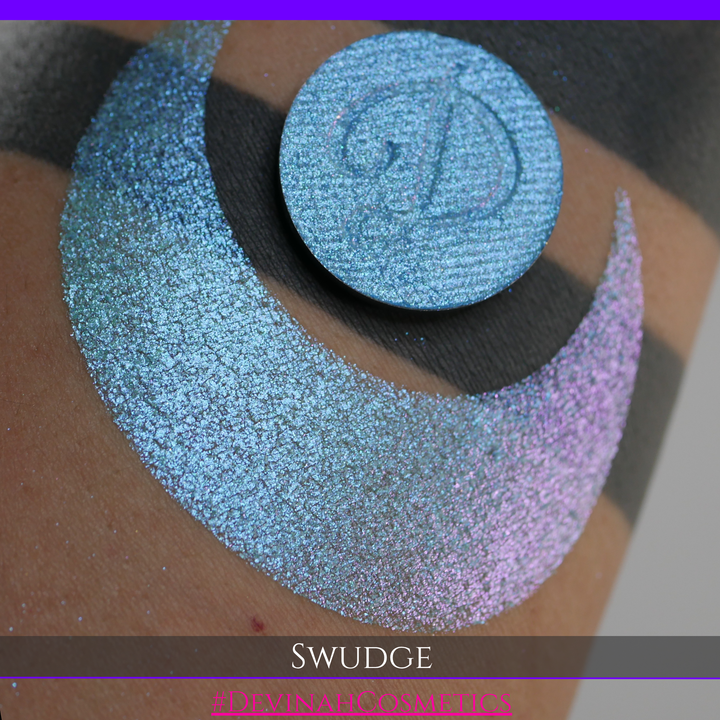 A round eyeshadow pan labeled 'SWUDGE' made by Devinah Cosmetics, placed over a large crescent moon swatch of the same shimmering shifty eyeshadow on skin. The eyeshadow has a duochrome multichrome shift between colors pink, teal, blue.