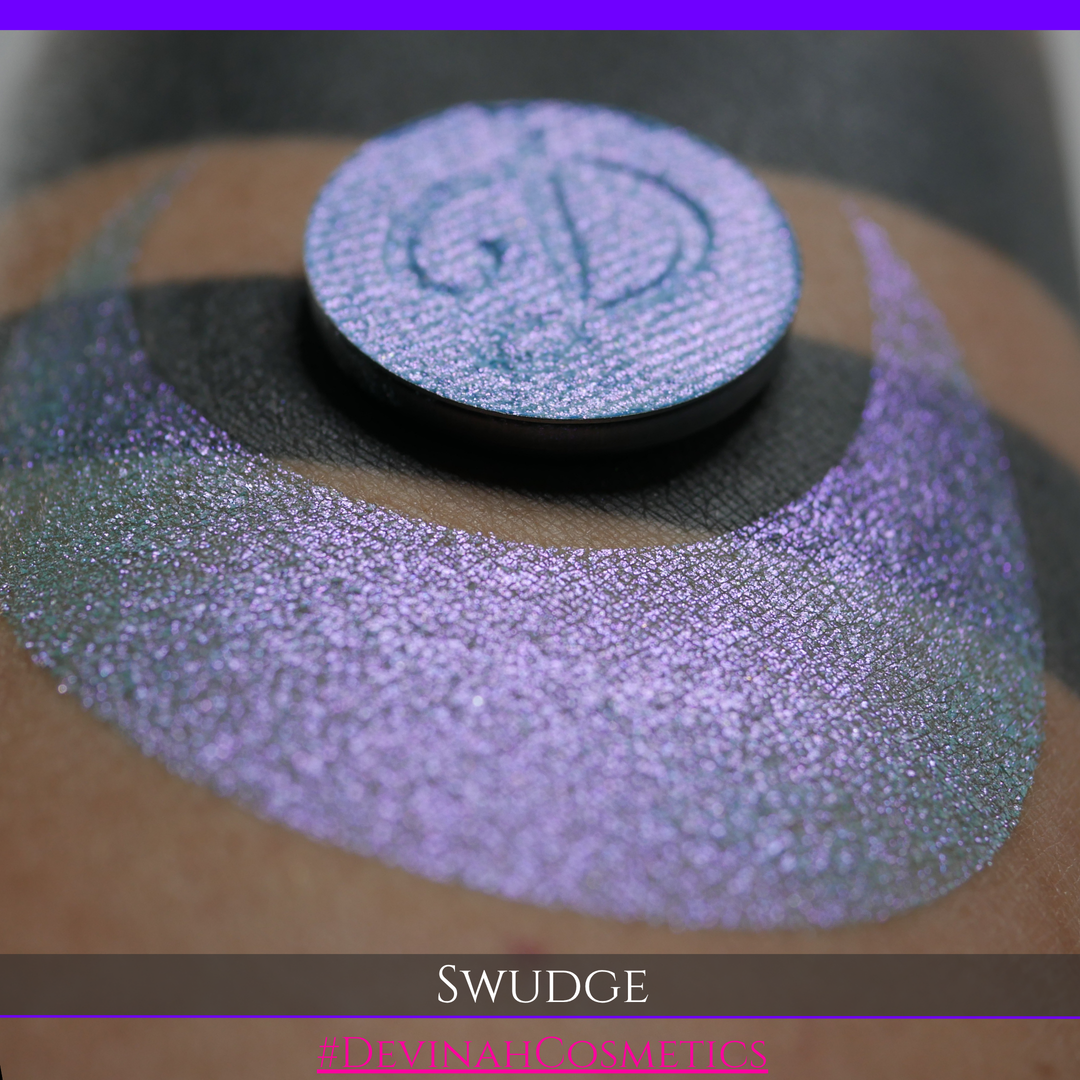 A round eyeshadow pan labeled 'SWUDGE' made by Devinah Cosmetics, placed over a large crescent moon swatch of the same shimmering shifty eyeshadow on skin. The eyeshadow has a duochrome multichrome shift between colors pink, teal, blue.
