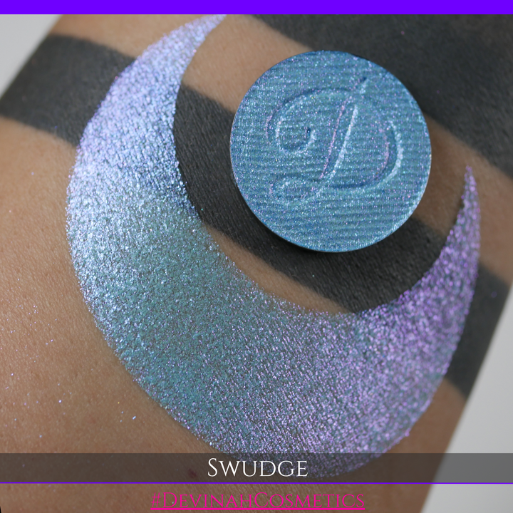 A round eyeshadow pan labeled 'SWUDGE' made by Devinah Cosmetics, placed over a large crescent moon swatch of the same shimmering shifty eyeshadow on skin. The eyeshadow has a duochrome multichrome shift between colors pink, teal, blue.