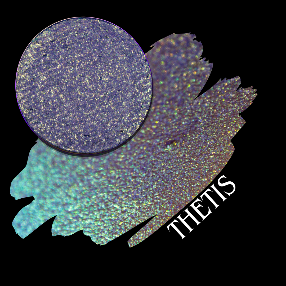 A cosmetic product swatch showing a circular eyeshadow of shimmering iridescent multichrome in a purple-to-teal gradient, labeled 'THETIS' on a black background made by Devinah Cosmetics.