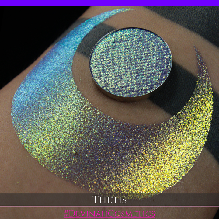 A close-up of a person's forearm showing a large iridescent crescent-shaped swatch in shimmering green, gold, and purple tones, with a round makeup eyeshadow pan of the same purple-toned glitter product placed on top. Text at the bottom reads 'THETIS' and '#DEVINAHCOSMETICS'.