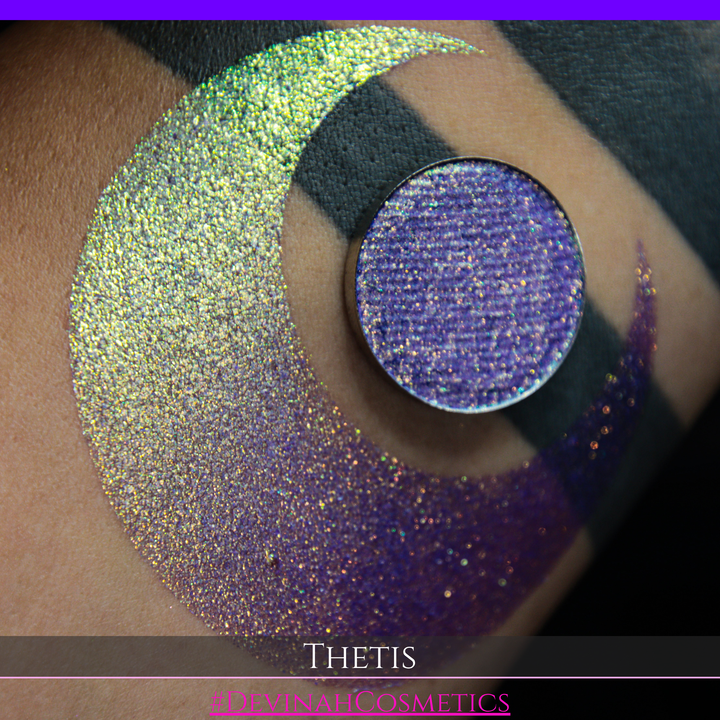 A close-up of a person's forearm showing a large iridescent crescent-shaped swatch in shimmering green, gold, and purple tones, with a round makeup eyeshadow pan of the same purple-toned glitter product placed on top. Text at the bottom reads 'THETIS' and '#DEVINAHCOSMETICS'.