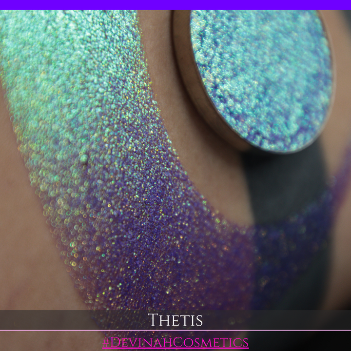 A close-up of a person's forearm showing a large iridescent crescent-shaped swatch in shimmering green, gold, and purple tones, with a round makeup eyeshadow pan of the same purple-toned glitter product placed on top. Text at the bottom reads 'THETIS' and '#DEVINAHCOSMETICS'.