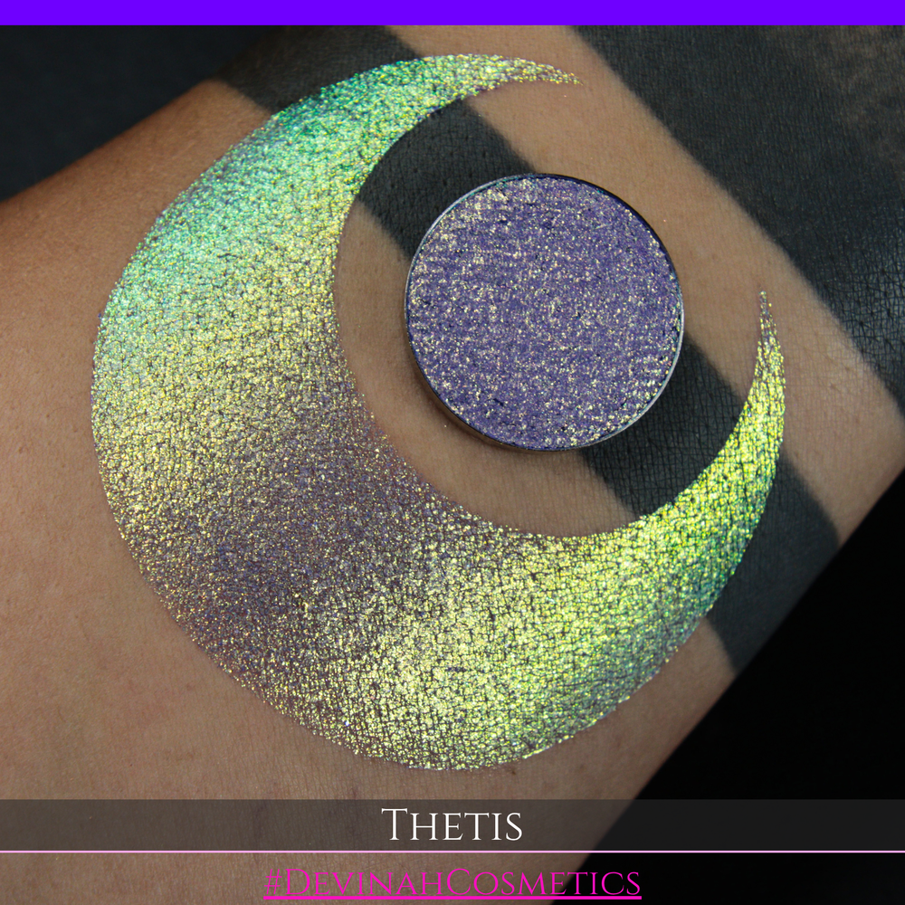 A close-up of a person's forearm showing a large iridescent crescent-shaped swatch in shimmering green, gold, and purple tones, with a round makeup eyeshadow pan of the same purple-toned glitter product placed on top. Text at the bottom reads 'THETIS' and '#DEVINAHCOSMETICS'.