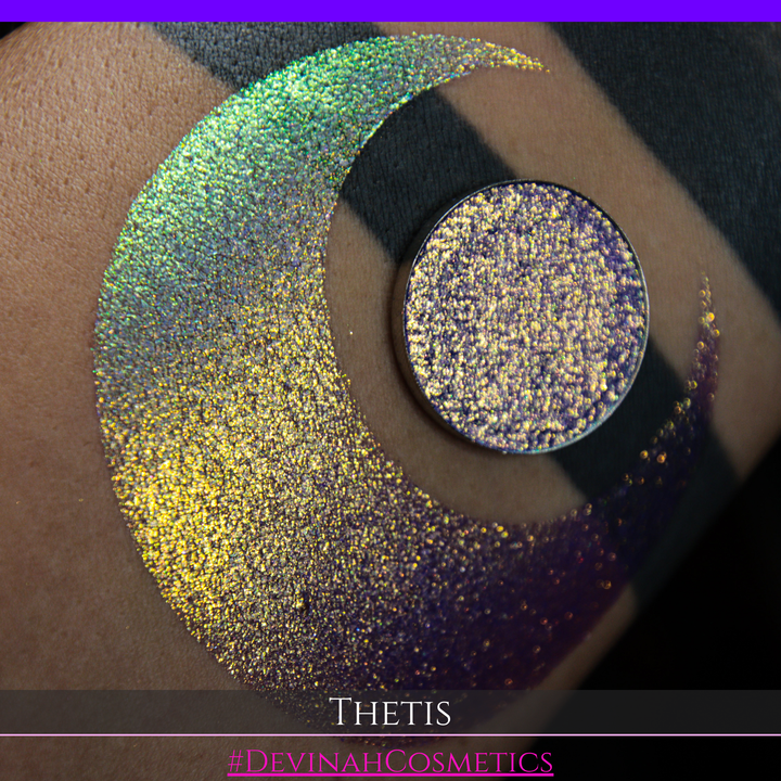 A close-up of a person's forearm showing a large iridescent crescent-shaped swatch in shimmering green, gold, and purple tones, with a round makeup eyeshadow pan of the same purple-toned glitter product placed on top. Text at the bottom reads 'THETIS' and '#DEVINAHCOSMETICS'.