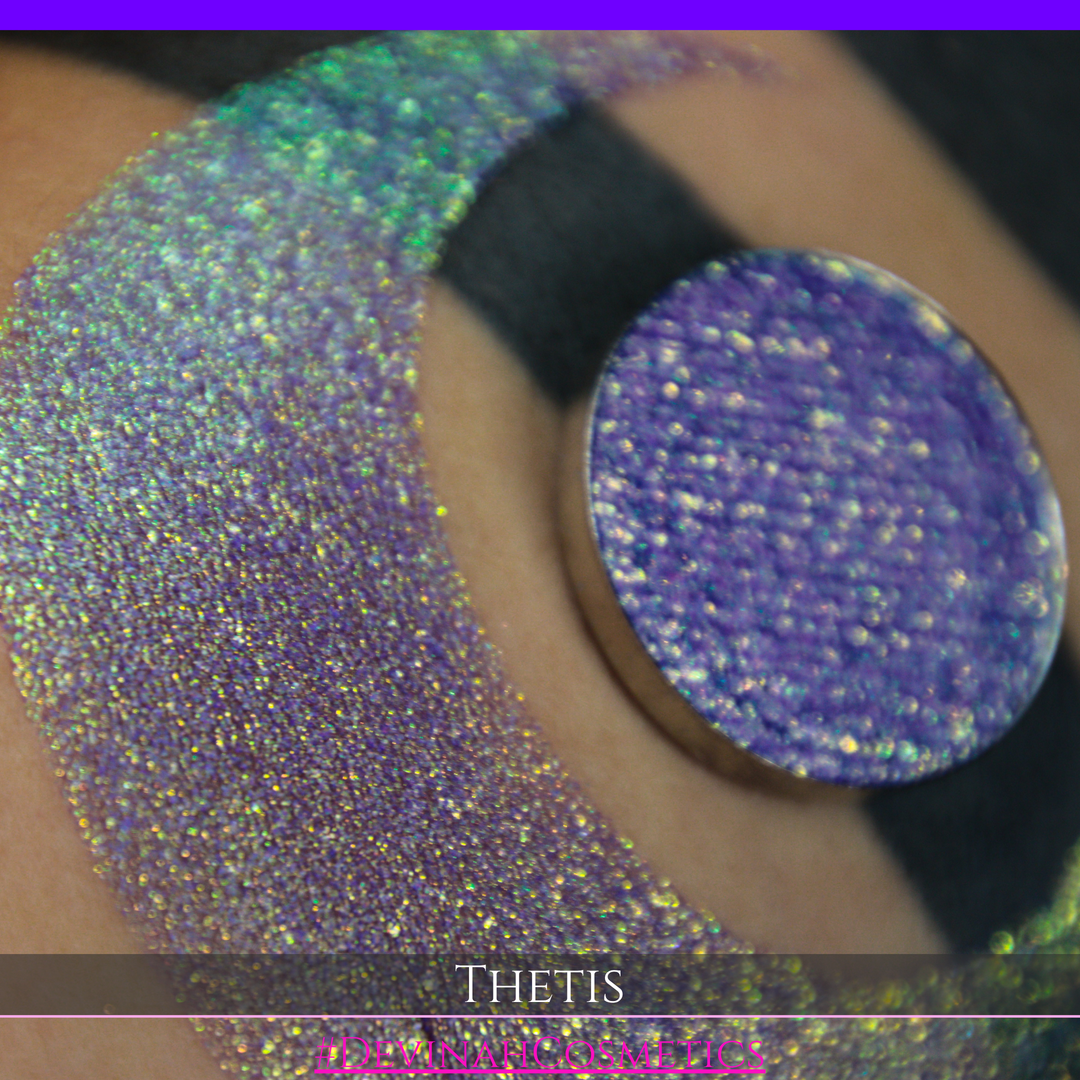 A close-up of a person's forearm showing a large iridescent crescent-shaped swatch in shimmering green, gold, and purple tones, with a round makeup eyeshadow pan of the same purple-toned glitter product placed on top. Text at the bottom reads 'THETIS' and '#DEVINAHCOSMETICS'.