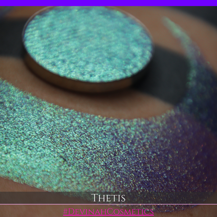 A close-up of a person's forearm showing a large iridescent crescent-shaped swatch in shimmering green, gold, and purple tones, with a round makeup eyeshadow pan of the same purple-toned glitter product placed on top. Text at the bottom reads 'THETIS' and '#DEVINAHCOSMETICS'.