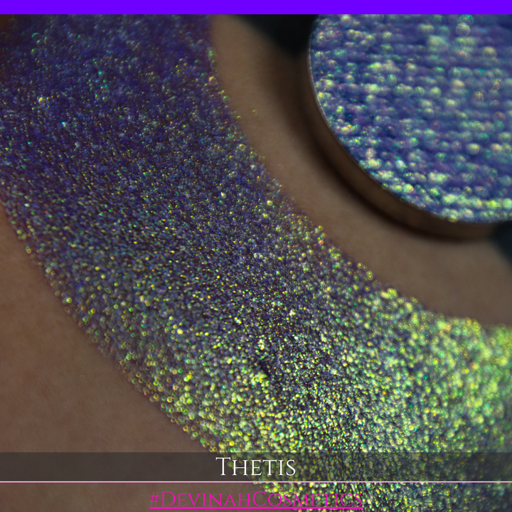 A close-up of a person's forearm showing a large iridescent crescent-shaped swatch in shimmering green, gold, and purple tones, with a round makeup eyeshadow pan of the same purple-toned glitter product placed on top. Text at the bottom reads 'THETIS' and '#DEVINAHCOSMETICS'.