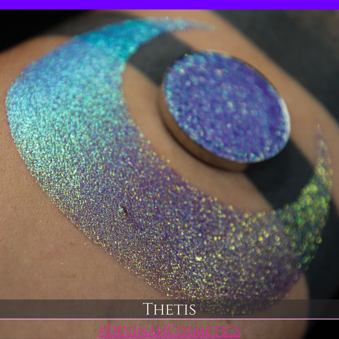 A close-up of a person's forearm showing a large iridescent crescent-shaped swatch in shimmering green, gold, and purple tones, with a round makeup eyeshadow pan of the same purple-toned glitter product placed on top. Text at the bottom reads 'THETIS' and '#DEVINAHCOSMETICS'.
