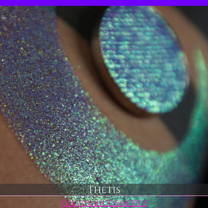A close-up of a person's forearm showing a large iridescent crescent-shaped swatch in shimmering green, gold, and purple tones, with a round makeup eyeshadow pan of the same purple-toned glitter product placed on top. Text at the bottom reads 'THETIS' and '#DEVINAHCOSMETICS'.