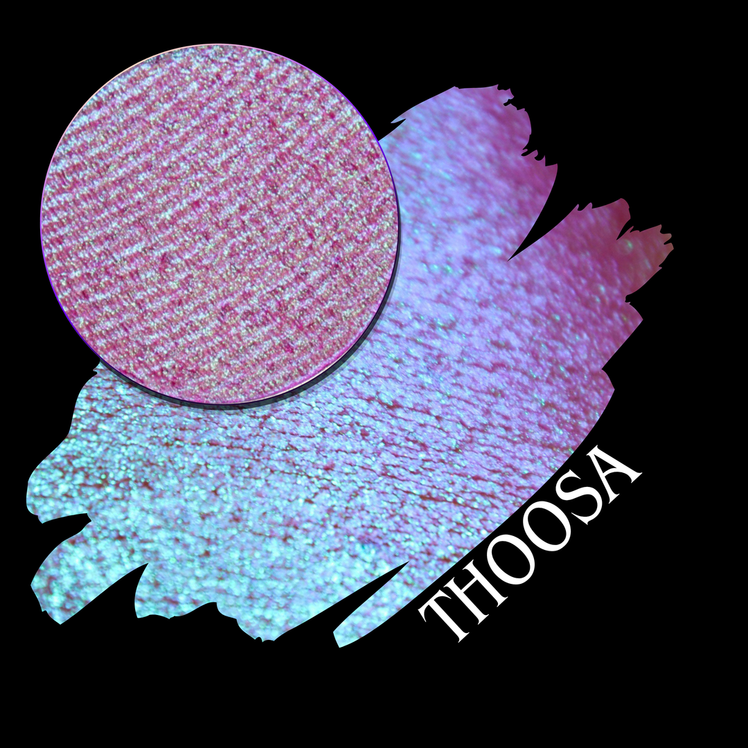 A round eyeshadow pan labeled 'THOOSA' made by Devinah Cosmetics, placed over a large crescent moon swatch of the same shimmering shifty eyeshadow on skin. The eyeshadow has a duochrome multichrome shift between colors blue, purple, green, pink.