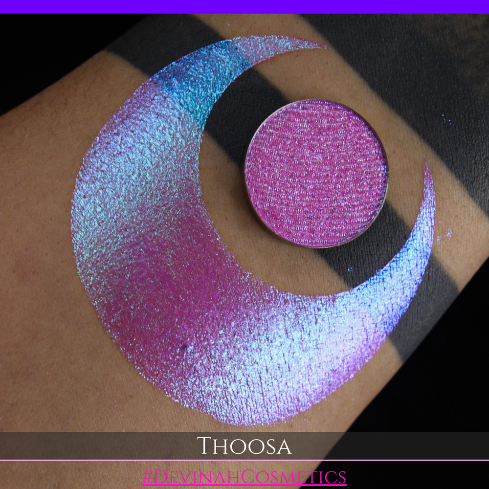 A round eyeshadow pan labeled 'THOOSA' made by Devinah Cosmetics, placed over a large crescent moon swatch of the same shimmering shifty eyeshadow on skin. The eyeshadow has a duochrome multichrome shift between colors blue, purple, green, pink.