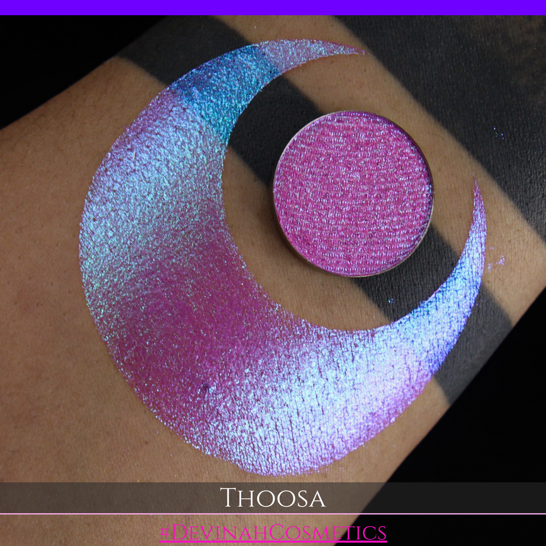 A round eyeshadow pan labeled 'THOOSA' made by Devinah Cosmetics, placed over a large crescent moon swatch of the same shimmering shifty eyeshadow on skin. The eyeshadow has a duochrome multichrome shift between colors blue, purple, green, pink.