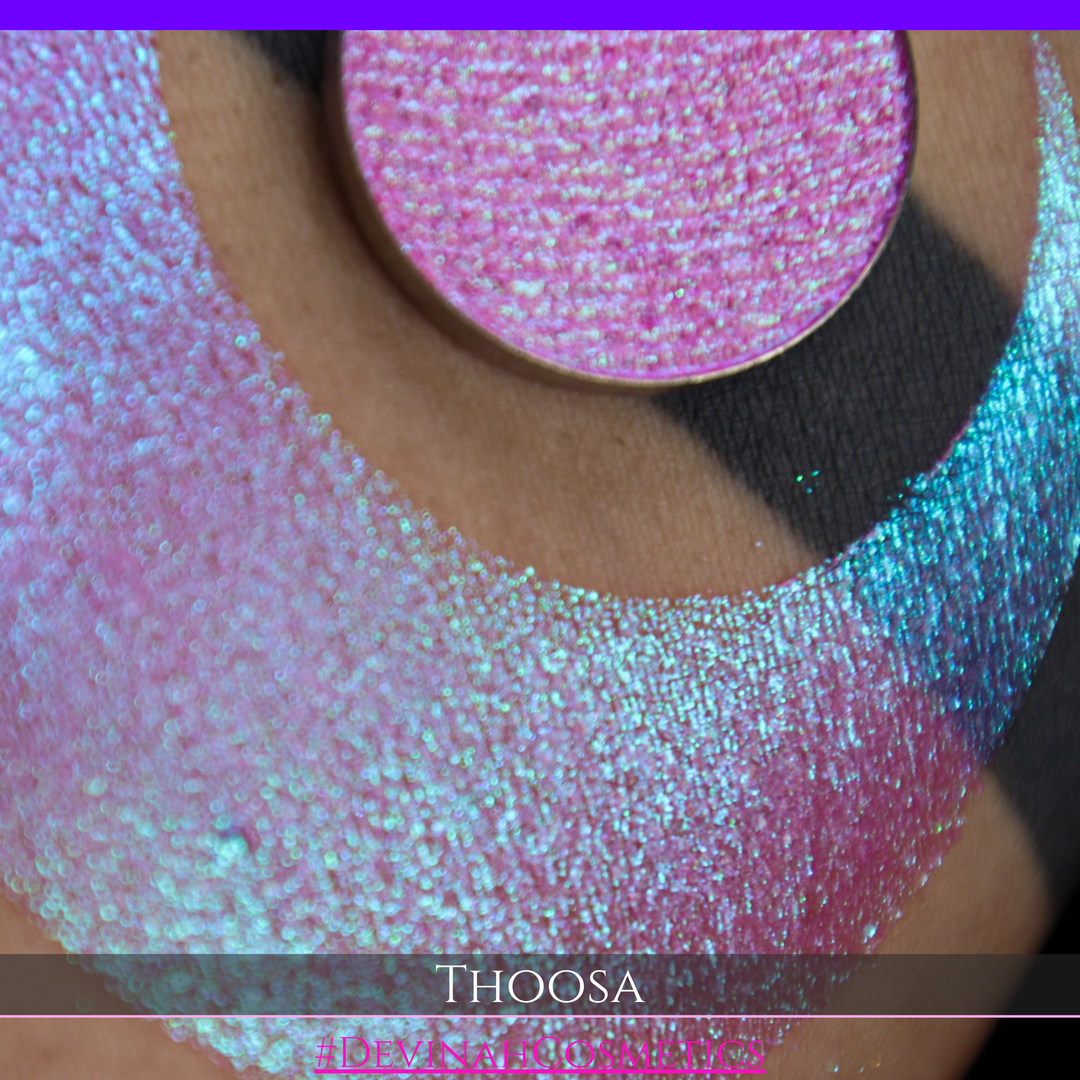 A round eyeshadow pan labeled 'THOOSA' made by Devinah Cosmetics, placed over a large crescent moon swatch of the same shimmering shifty eyeshadow on skin. The eyeshadow has a duochrome multichrome shift between colors blue, purple, green, pink.