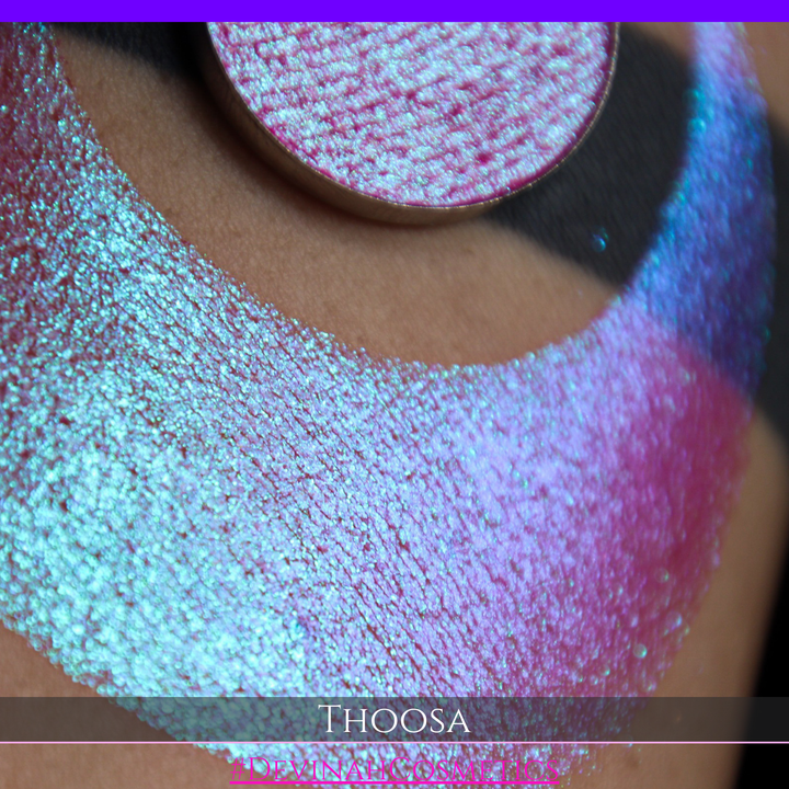 A round eyeshadow pan labeled 'THOOSA' made by Devinah Cosmetics, placed over a large crescent moon swatch of the same shimmering shifty eyeshadow on skin. The eyeshadow has a duochrome multichrome shift between colors blue, purple, green, pink.