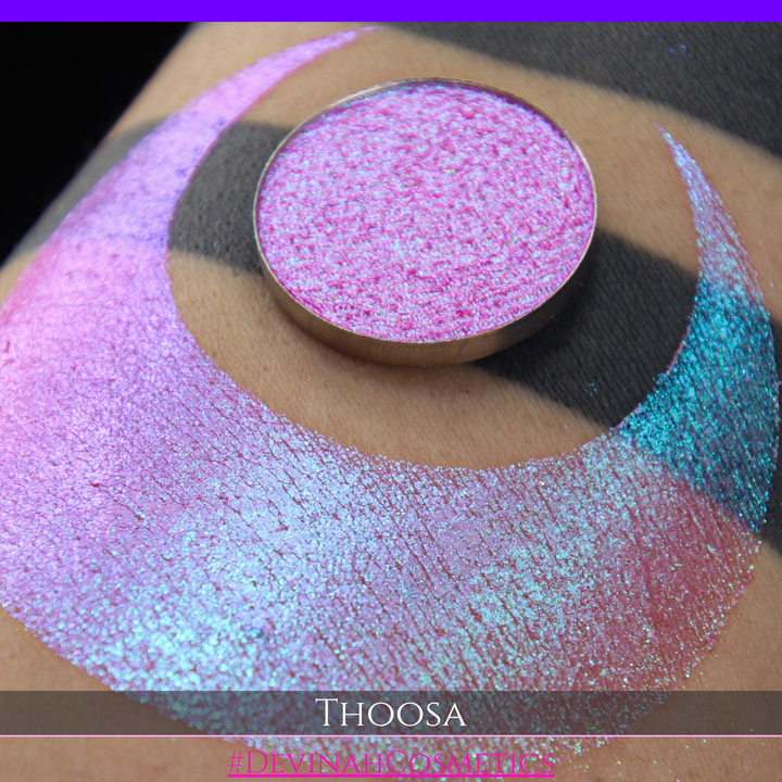 A round eyeshadow pan labeled 'THOOSA' made by Devinah Cosmetics, placed over a large crescent moon swatch of the same shimmering shifty eyeshadow on skin. The eyeshadow has a duochrome multichrome shift between colors blue, purple, green, pink.