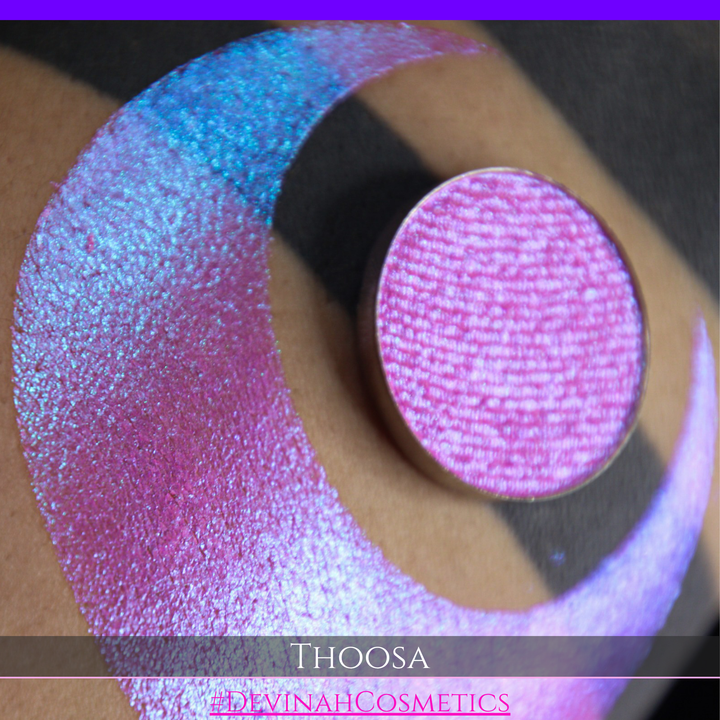 A round eyeshadow pan labeled 'THOOSA' made by Devinah Cosmetics, placed over a large crescent moon swatch of the same shimmering shifty eyeshadow on skin. The eyeshadow has a duochrome multichrome shift between colors blue, purple, green, pink.